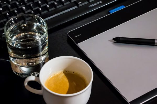 Office desk setup with a coffee cup, glass of water, tablet, pen, and keyboard.